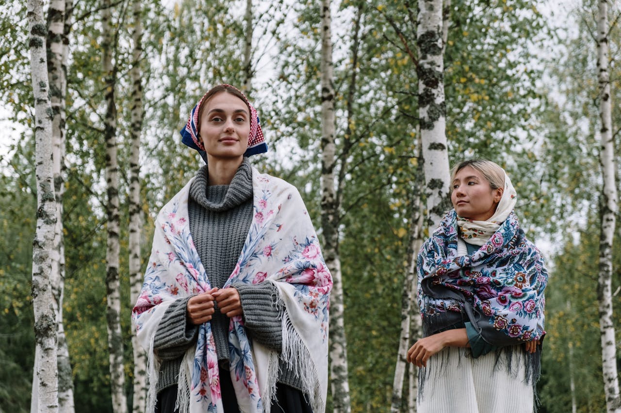 hero-img-01 Two women in knitted sweaters and traditional scarves stand together in a birch forest.