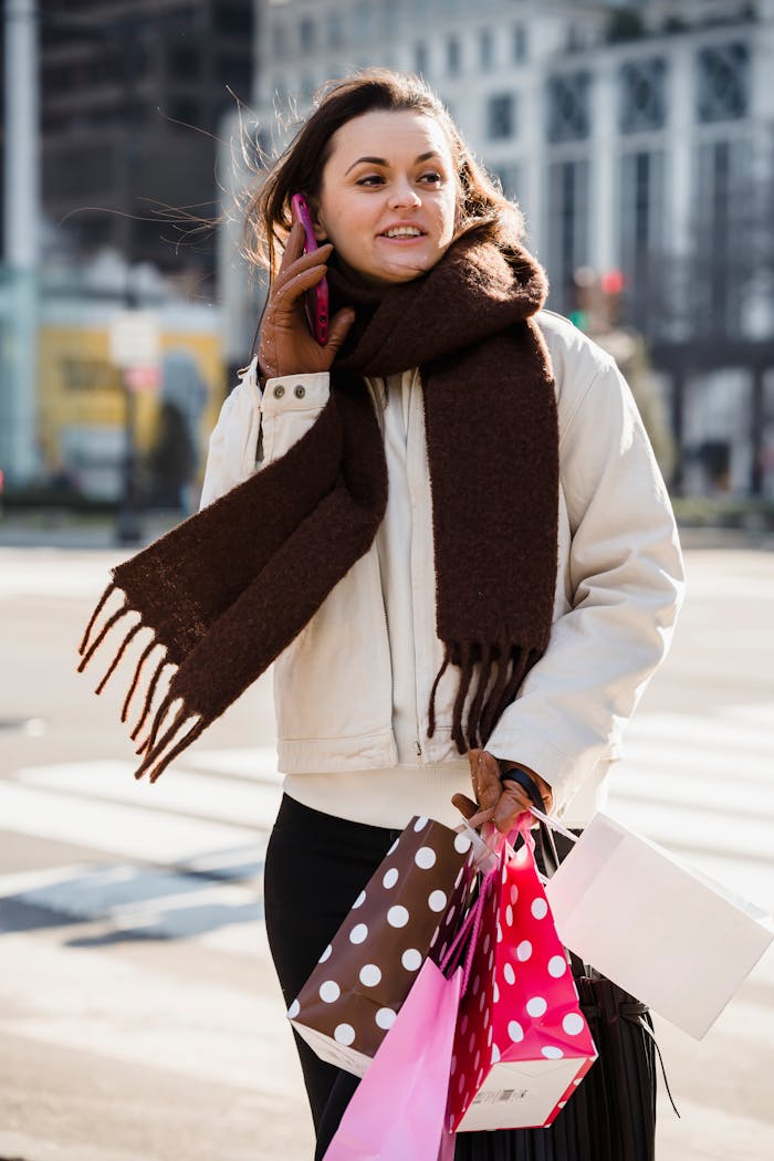 about-01 Happy young female in brown scarf smiling and looking away while talking on mobile phone on blurred background of street